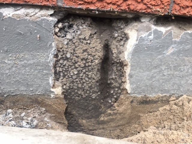 Husbetongssockel med skadat område där färg och betong lossnat, exponerande brun sand/jord.