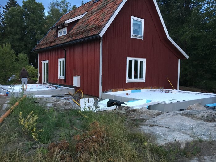 Grundarbeten för utbyggnad vid röd stuga med isolering och armerade kantbalkar synliga i skymningen.