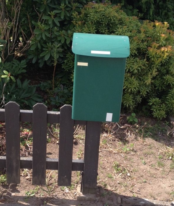 Grön postlåda monterad på ett nybytt mörkt trädgårdsstaket, omgiven av växtlighet.