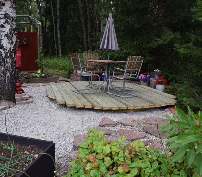 Uteplats med trägolv och trädgårdsmöbler inramad av grönska, grus och en röd utedusch.