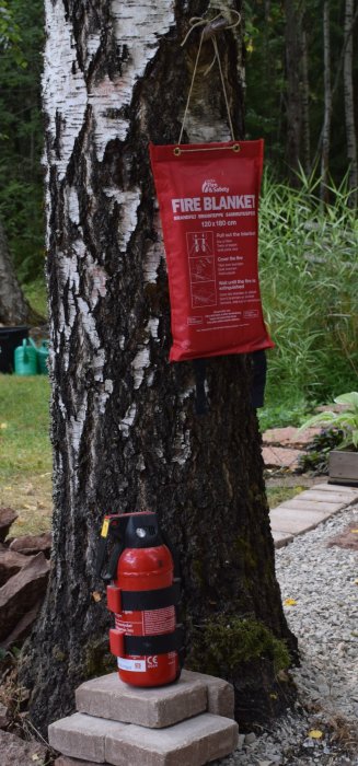 Brandfilt och brandsläckare placerade vid en björk på en grusbelagd uteplats omringad av skog.