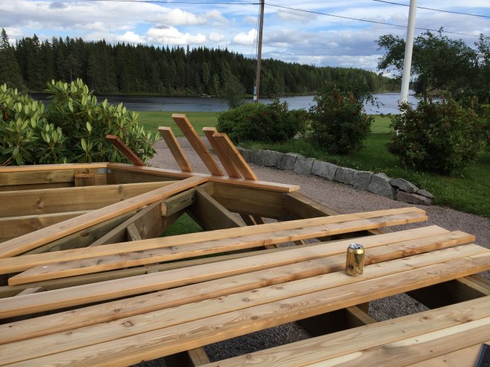 Skelett till en träaltan under konstruktion med en öppen ölburk, med utsikt över en sjö och grönska.