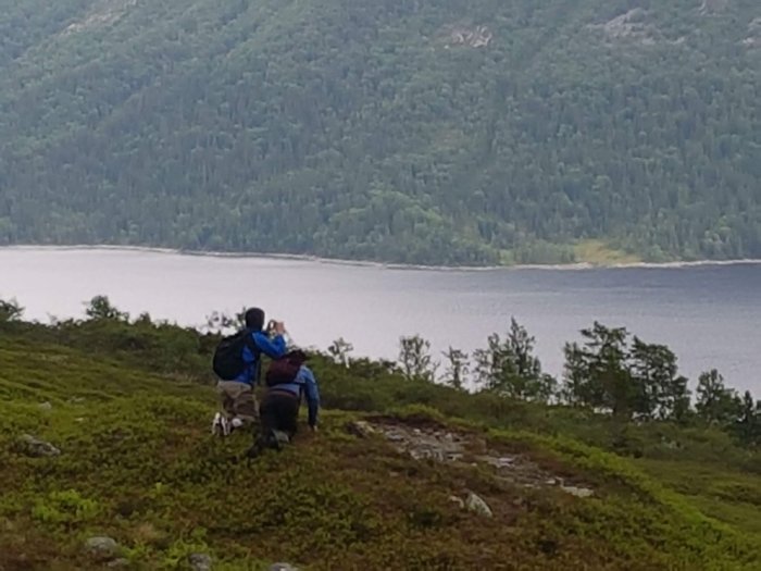 Två personer och en hund vilar på en kulle med utsikt över en sjö och berg i dis.