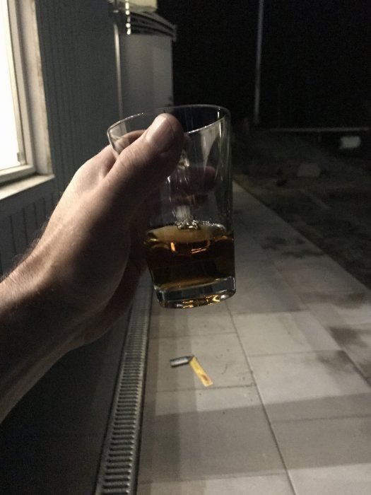 En hand som håller ett glas med en mörk dryck på en terrass om kvällen.