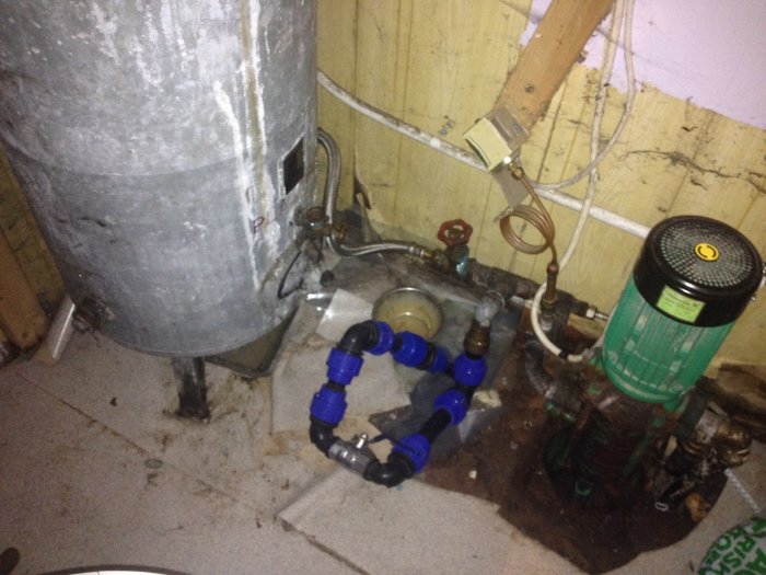 Ett hydrofortank och en grön dränkbar pump i ett pannrum med synliga rörkopplingar och kablar.