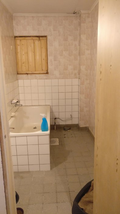 Äldre badrum med slitna klinkergolv och vit kaklad vägg, med borttaget badkar redo för renovering.