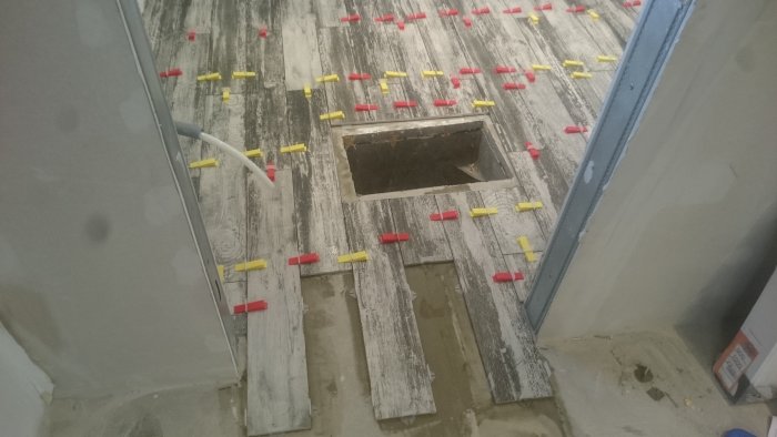Renoveringsarbete med läggning av gråa träimitationsplattor och avståndskilar runt ett golvbrunnshål.