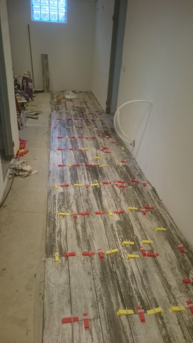 Ojämna plattor med röda och gula distanser på ett ofärdigt golv i en korridor under renovering.