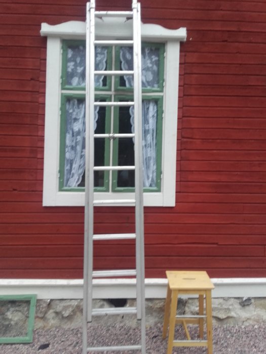 Nyinstallerat blindfönster på röd trävägg med vit kant och en stege framför samt en liten pall till höger.