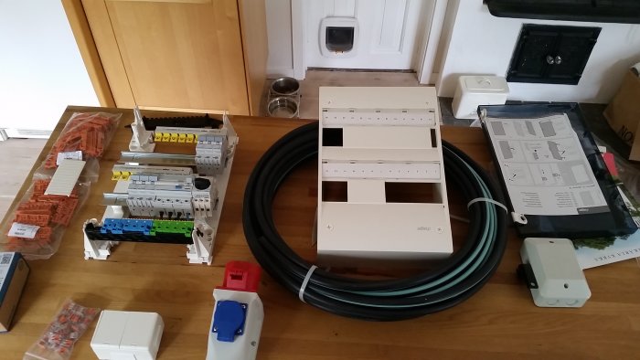 Elmaterial som normcentral, säkringar, kablar och vägguttag spridda på ett trägolv redo för installation.