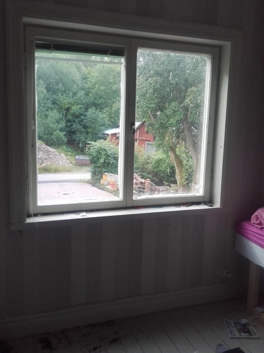 Nyinstallerat fönster i ett renoveringsprojekt, med utsikt över en trädgård, delvis tapetserad vägg och oslipat trägolv.