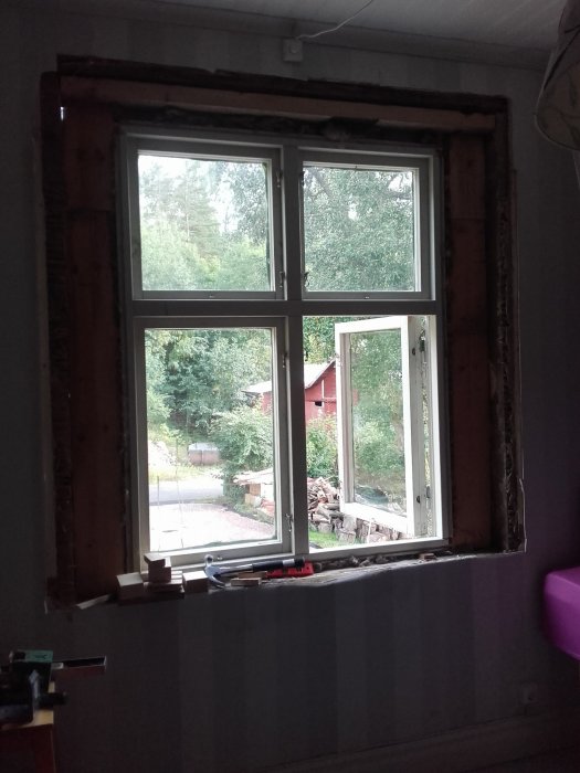 Nytt fönster med karm installerat i en husvägg under renovering, synliga verktyg och omgivande natur.
