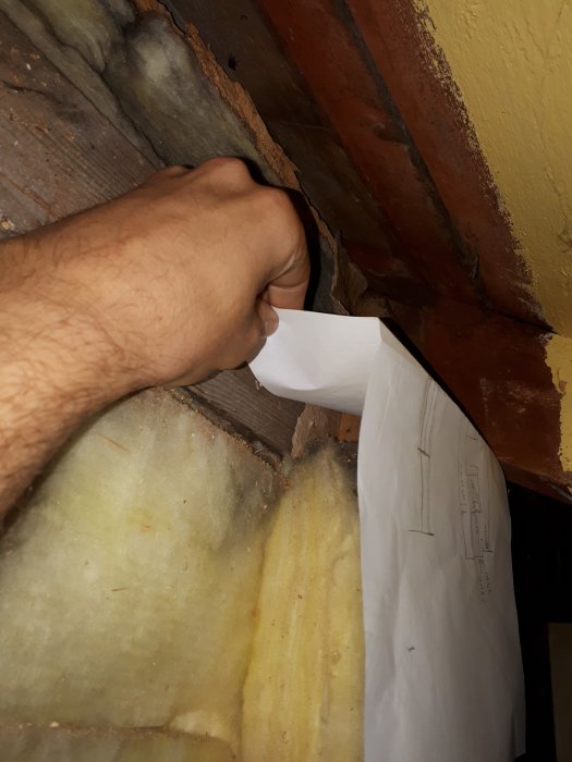 Hand som håller vit vindduk mot ett undertak med befintlig gul isolering och trästrukturer.