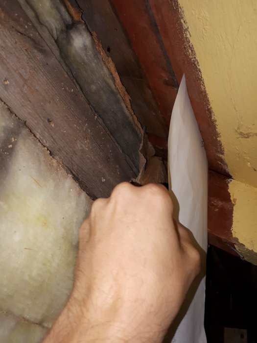 Hand som fäster vindduk mot ett trä undertak vid isolering med synlig gammal isolering.
