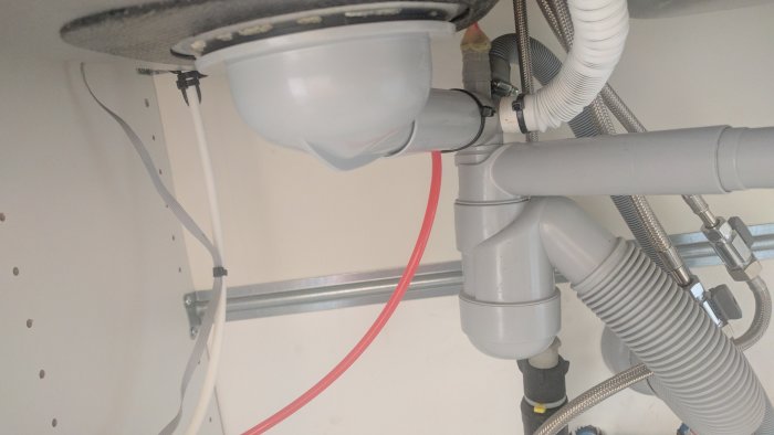 Rör och slangar under diskho inklusive avlopp, vattenledningar och en röd slang.