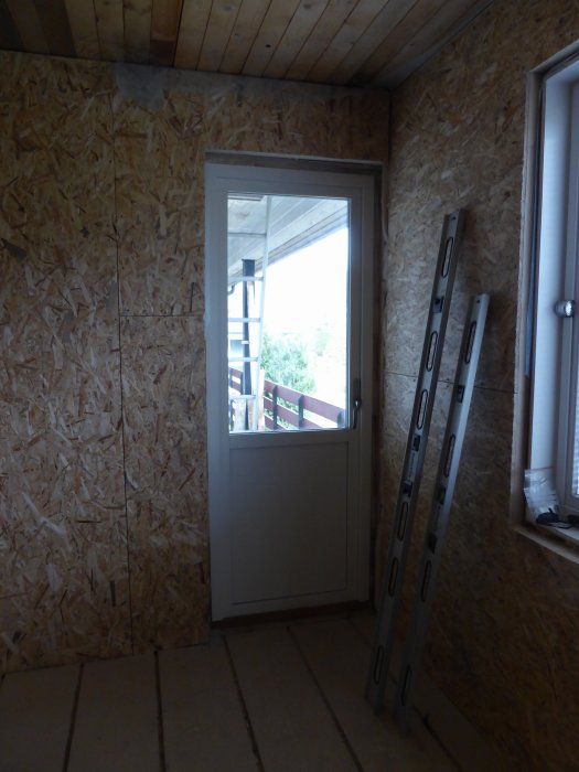 Pågående renovering i stort sovrum med OSB-väggar, golvskivor och en nyinstallerad balkongdörr, samt en lutande bygglaser.