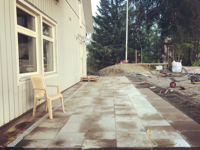 Stensatt uteplats under konstruktion med nyfogade plattor och byggmaterial nära vit husvägg.