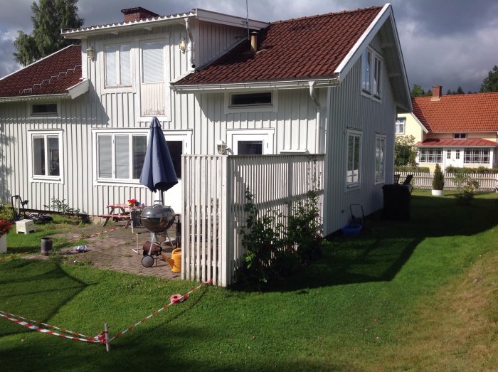 Trädgård med uppmätt område för trall framför vitt hus med gräsmatta och grill.