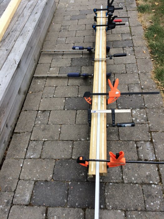 Kluvna träbitar som vändlimmats med PU-lim och hålls samman av flera spännklämmor på en stenlagd mark.