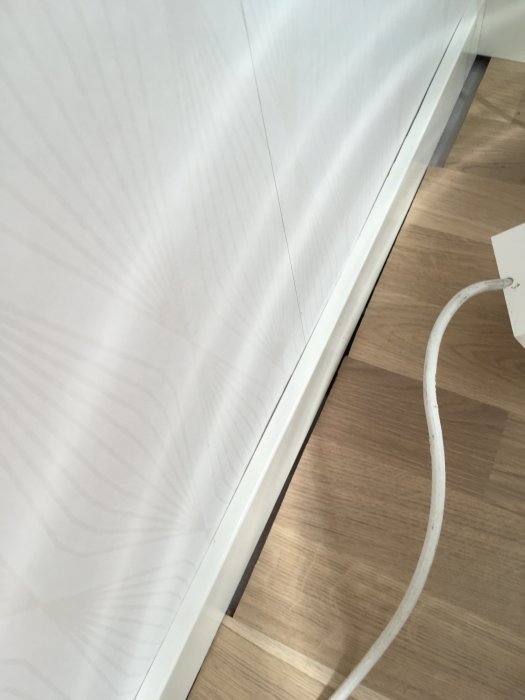 Dåligt lagt trägolv med synlig glipa vid vit vägg och golvlister, kabel ligger på golvet.