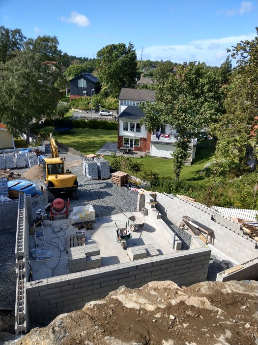 Byggarbetsplats med grävmaskin, materialhögar och pågående konstruktion av grå blockväggar.