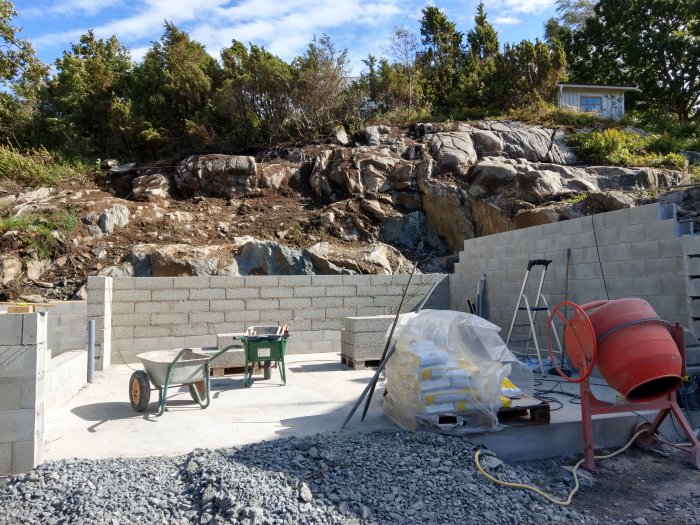 Byggplats med gråsten, betongblockvägg under konstruktion, skottkärra, betongblandare och byggmaterial.