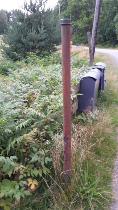 Rostig metallpinne som kan vara en gammal tomtpinne, vid en grusväg med grönska och brevlådor i bakgrunden.