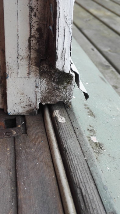 Sliten altandörrtröskel med skadade fönsterlister och tecken på fuktintrång vid golvet.
