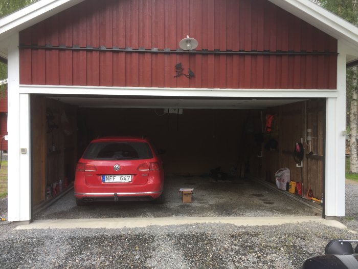 Öppet kallgarage på grusunderlag med röd VW-bil parkerad inuti, väggar av trä, och lecablock synliga vid grund.