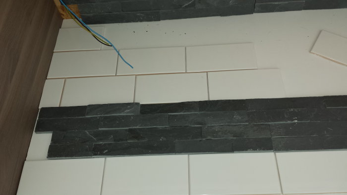 Testläggning av vit kakel och mörk dekorsten utan fog, mot en vägg under renovering.