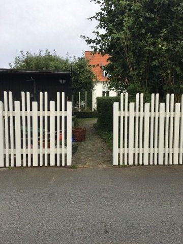 Vitt staket med en synlig glipa mellan staketsektionerna vid en uppfart med grind framför en trädgård.