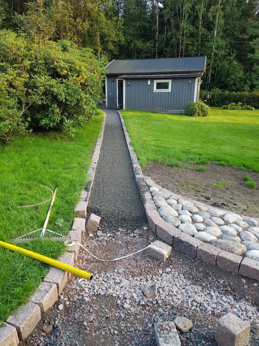 En nygrusad gångväg bredvid en stenmur med pågående arbete framför ett grått hus med grönt gräs.