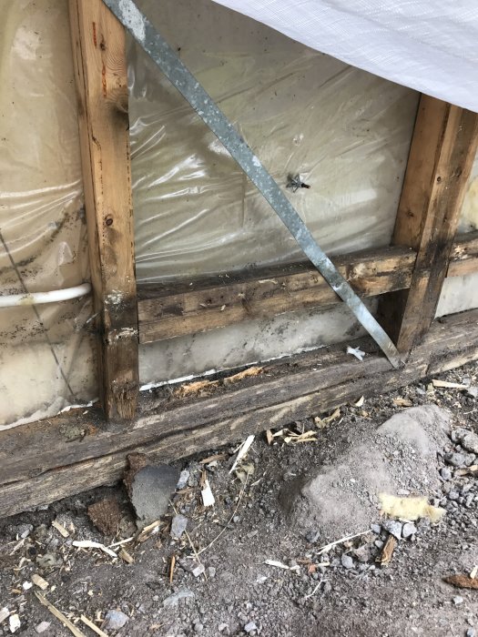 Detaljbild av ett byggprojekt som visar isolering och träreglar bakom en presenning och tecken på fukt i underliggande trä.