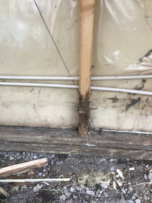 Skadad trästolpe med rötskador vid husgrund med isolering och elrör.