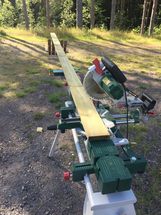 Ett långt trästycke som kapas med kap- och geringssåg på en sågbock i utomhusmiljö med skog och gräsmatta i bakgrunden.