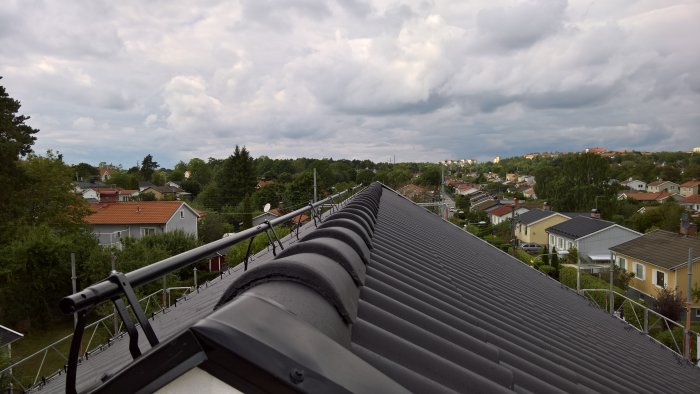 Utsikt från ett tak med nockband på plats och påbörjad fasadbelysning, över ett villaområde med molnig himmel.