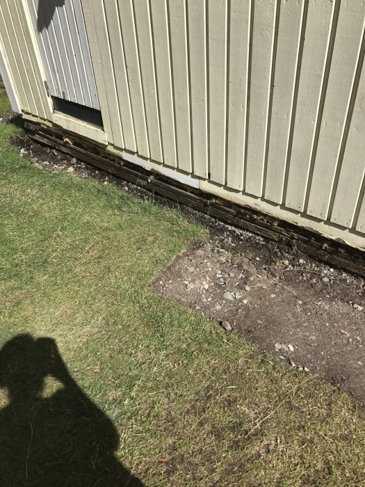 Husets hörn med avlägsnad panel visar en träsyll som ligger på marken, intill en grävd yta under reparation.
