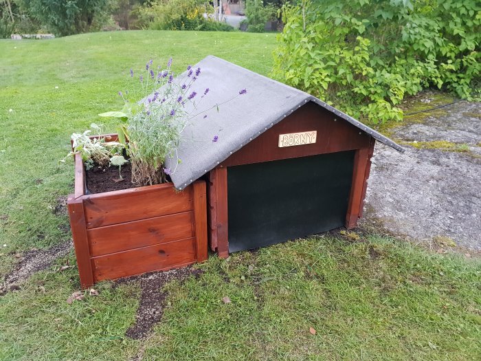 Litet trähus med skylt "BÖRNY", integrerad blomlåda med växter, på en grön gräsmatta.