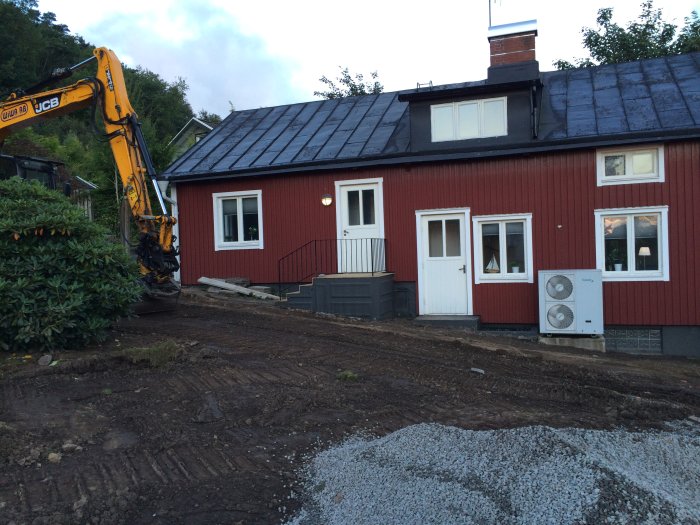 Röd stuga med ett slitit galvaniserat plåttak och flagnande färg, en grävmaskin till vänster och grushögar i förgrunden.