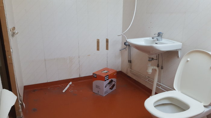 Ett halvrenoverat badrum med kakelväggar, handfat, toalett och en kartong på golvet.