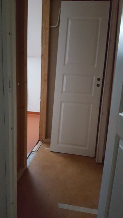 En öppen vit dörr som leder till ett rum under renovering med synliga träreglar och kablar.