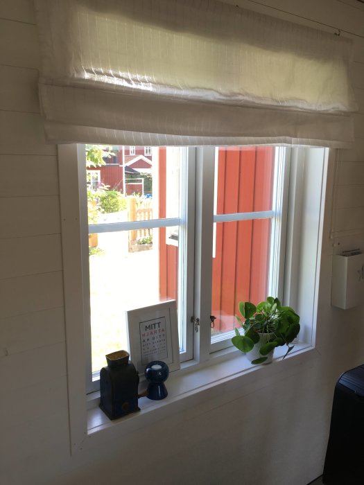 Ett vitmålat fönster med vita fönsterfoder, en vit rullgardin uppe och inredningsobjekt på fönsterbrädan.