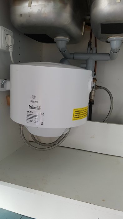 Nyinstallerad varmvattenberedare under diskbänk med kopplade vattenledningar och avloppsslang.