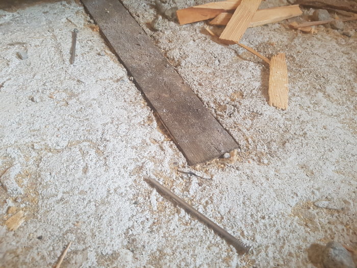 Uppbruten betonggolv med synliga träspill, en träregel och spikar, indikerande renoveringsarbete.