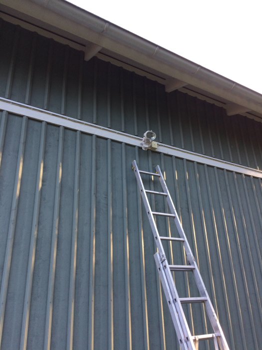 Ny utomhuslampa med sensor installerad högt upp på en husvägg, stege nedanför.