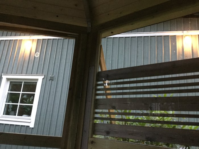 Kvällsbelysning på en uteplats med trästaket, växtlighet och del av ett hus med fönster.