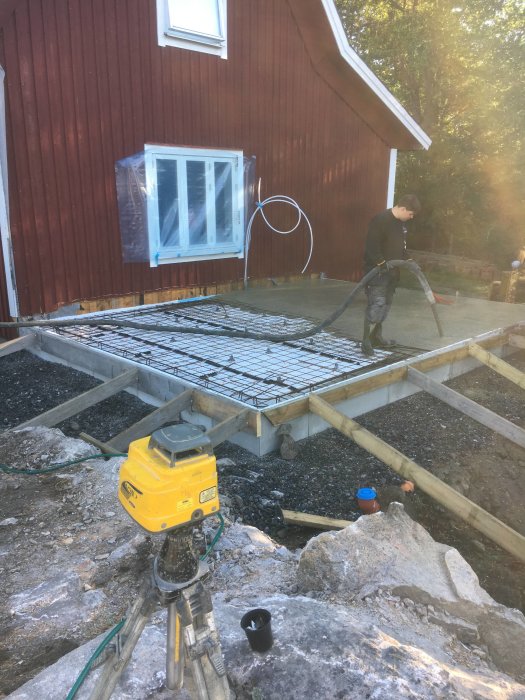 Person som gjuter betong på armeringsjärn nära en röd trähusfasad.