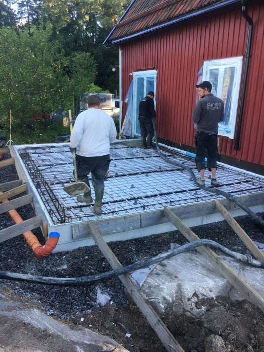 Personer förbereder gjutning av betong på armeringsnät vid byggprojekt nära röd stuga.