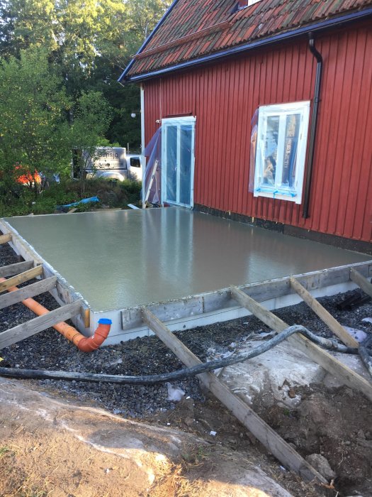 Nyligen gjuten betongplatta vid röd trähusgrund med träformar och grusunderlag.