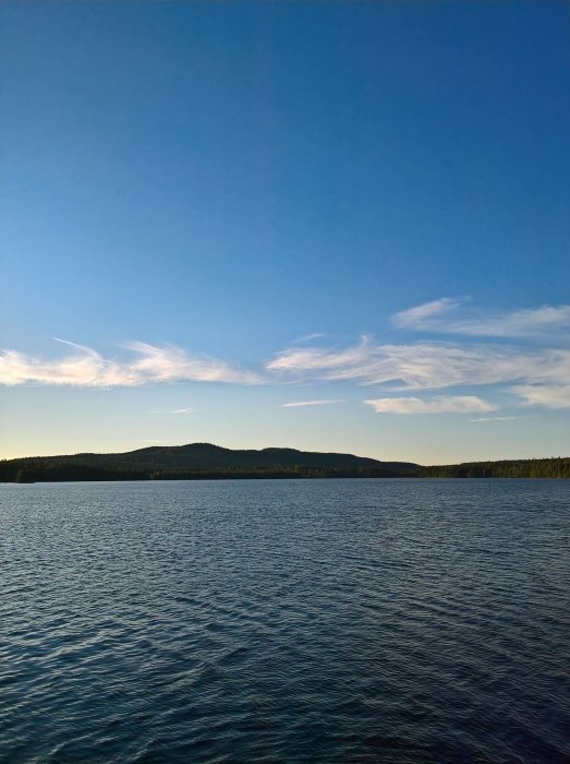 Vy över en lugn badplats med stilla vatten och skogklädda kullar i bakgrunden mot en klar himmel.